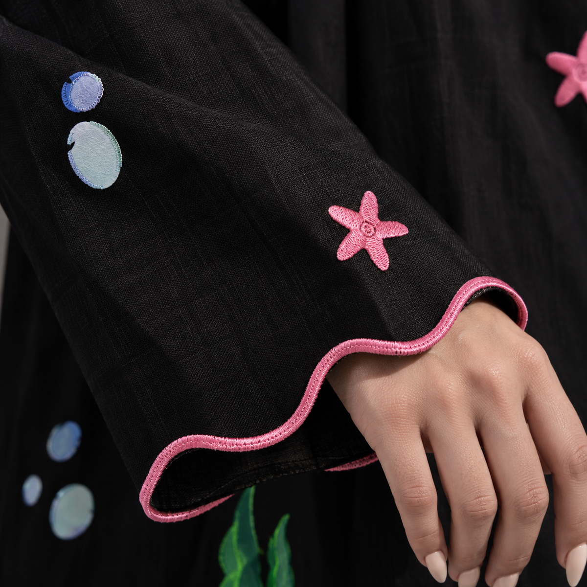 Close-up of a black garment sleeve with scalloped pink trim and embroidered pink starfish and blue bubble motifs, shown on a person's hand.